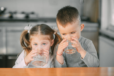 Two young children in a kitchen drinking clear tap water tested with a Kleer Blue Water testing kit for home.