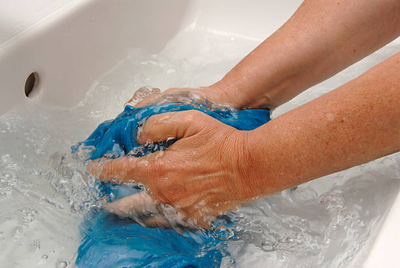 Hands washing a blue garment in a sink with tap water tested for iron, hardness, and staining with a water test kit.
